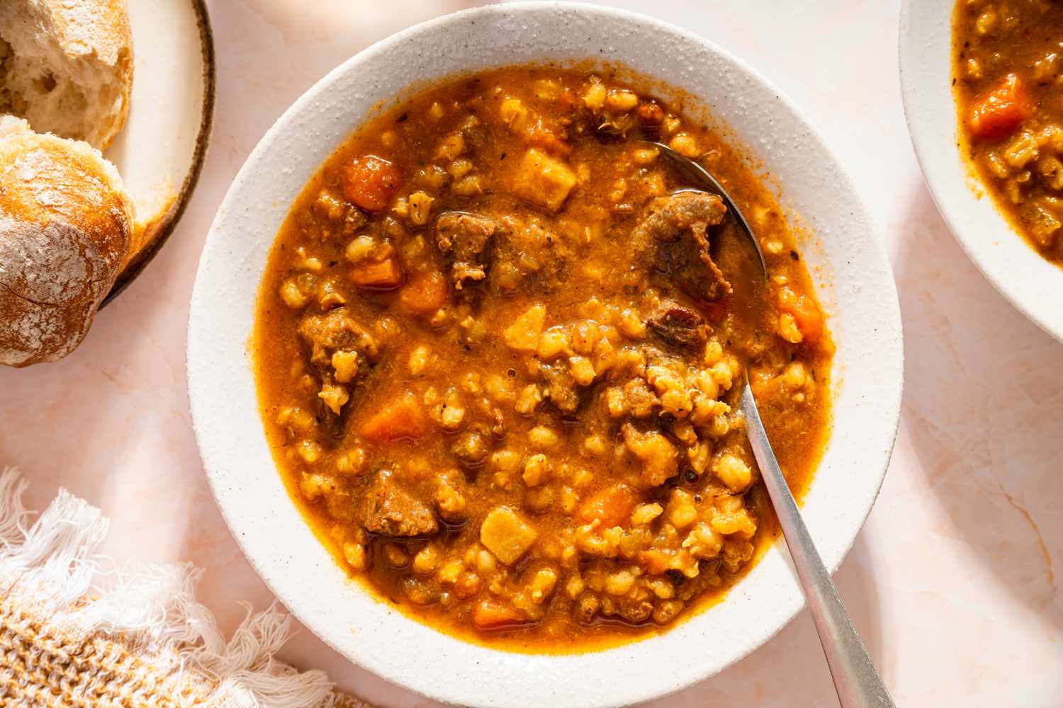 A bowl of beef barley soup with a spoon served with bread on the side