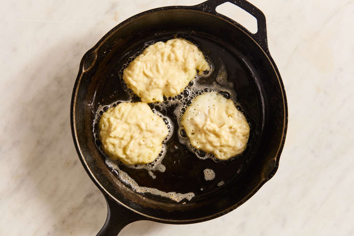 Overhead view of a cast iron skillet of three potato pancakes cooking from Boxty (Irish Potato Pancakes) recipe