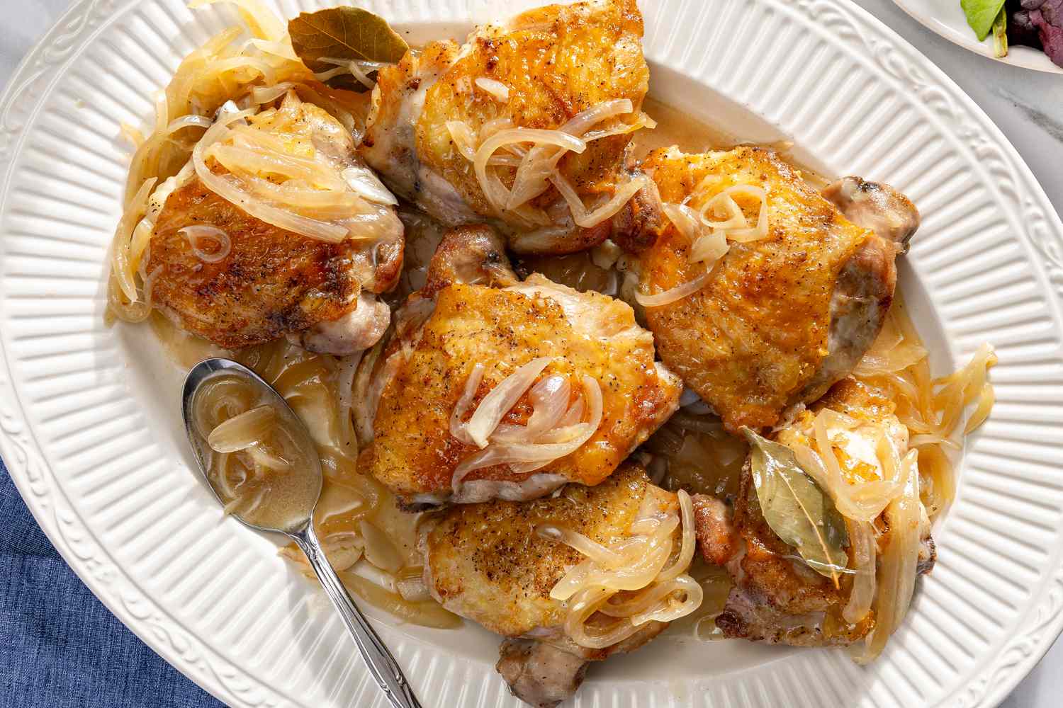 Overhead view of a white platter of six wine roasted chicken thighs topped with onions and bay leaves along with a spoon