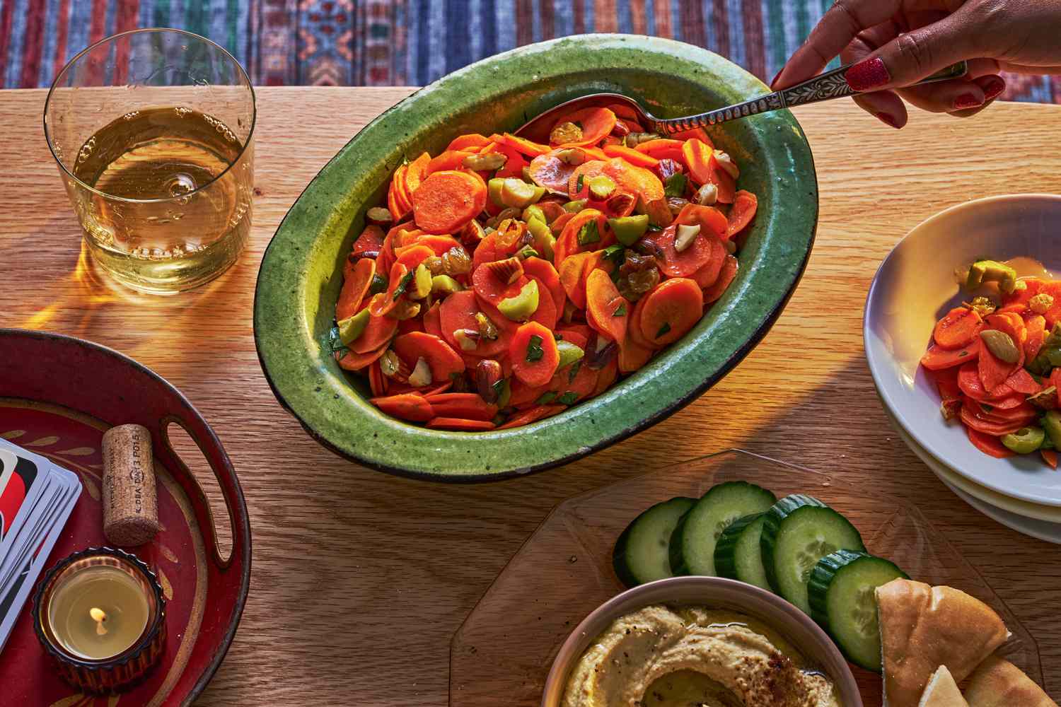 A bowl of carrot salad on a coffee table, alongside hummus with cucumbers and pita and a glass of wine