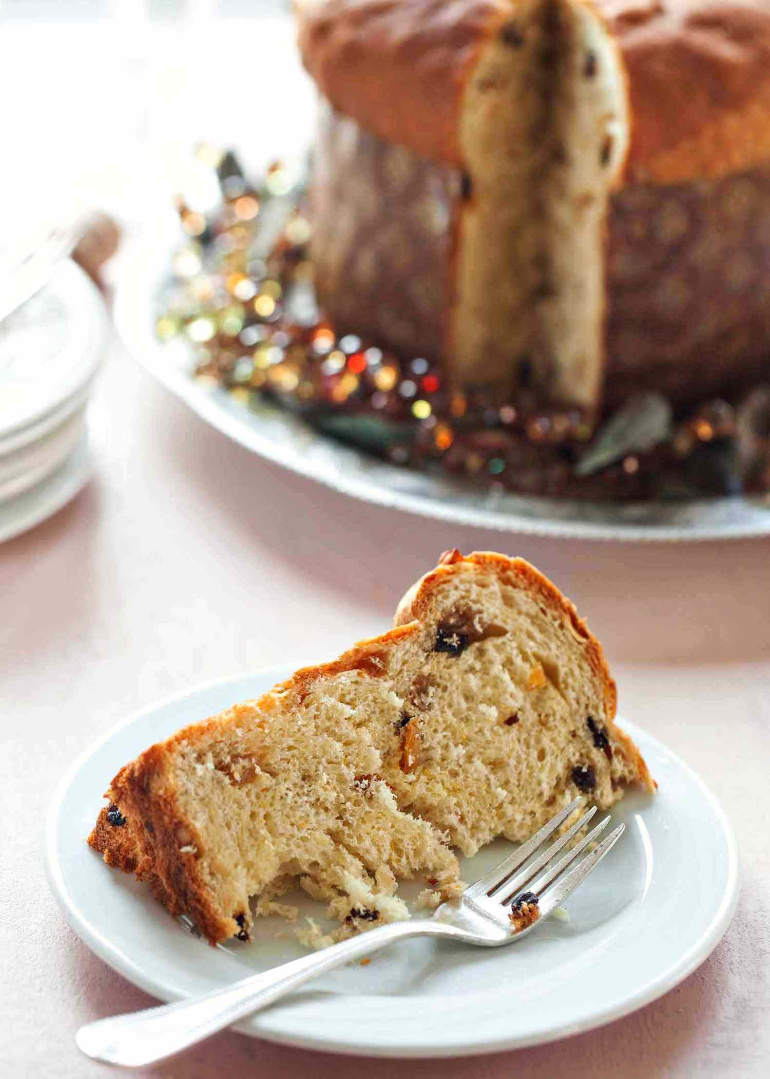 A slice of Panettone, Italian Chirstmas Bread, on a plate with a fork while the rest of the loaf is in the background.