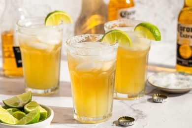 Three glasses of bebidas garnished with lime wedges with bottles and lime wedges in the background