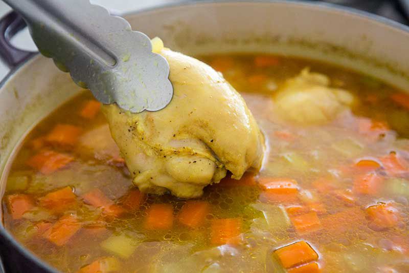 Removing cooked chicken thigh with tongs from a pot of soup 