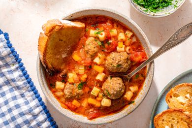 Overhead view of a bowl of Italian meatball soup with a slice of bread and a spoon next to a plate of bread and bowl of parsley