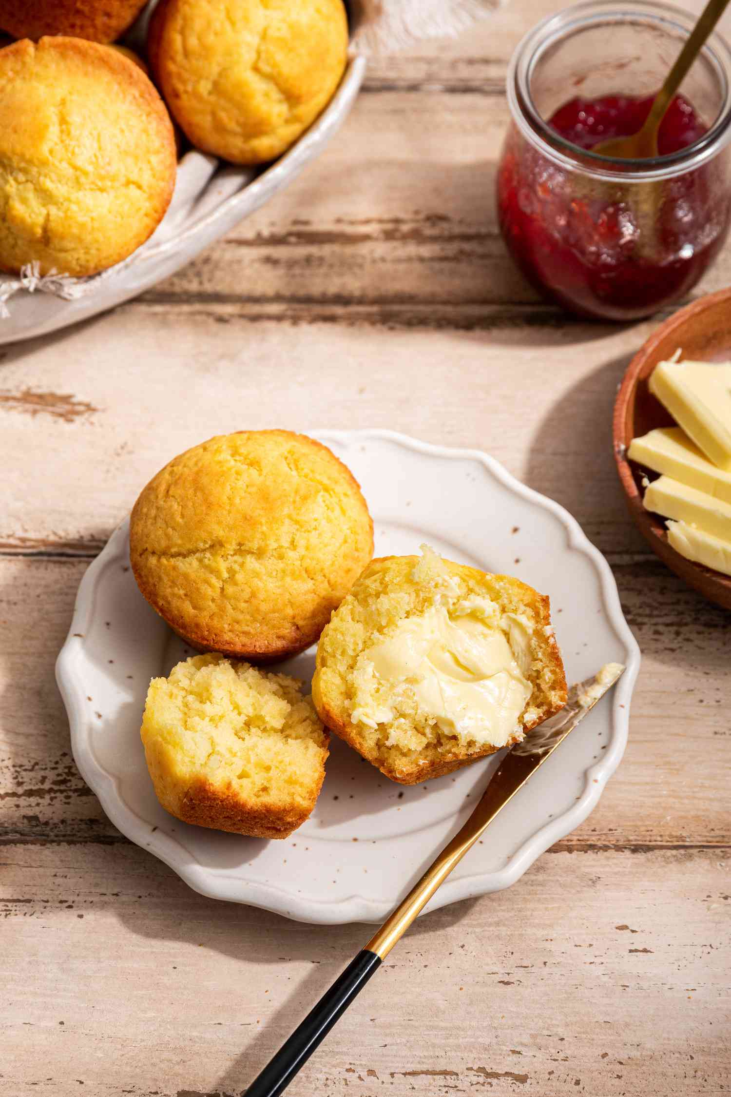 A nothin' muffin cut in half and smeared with butter on a small plate next to another nothin' muffin, and in the surrondings, a bowl with more muffins, a small bowl of butter, and a jar of jam