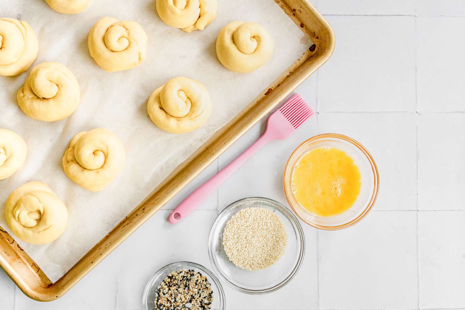 Challah Rolls on Baking Sheet Next to Three Bowls (One with an Egg Wash, Another with Sesame Seeds, and the Last with a Spice Blend)