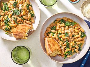 Overhead view of a bowl of beans and greens with a chunk of bread all next to a spoon and resting on a blue cloth napkin