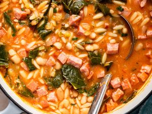 Pot of soup with beans orzo diced ham and greens served with a ladle and spoon