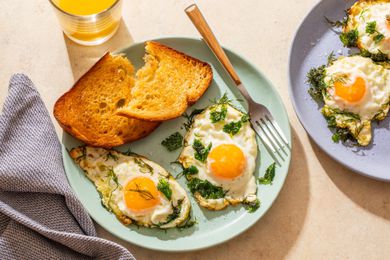 Fried eggs and toat on a plate at a table setting with a glass of orange juice, another plate of eggs, and a kitchen towel