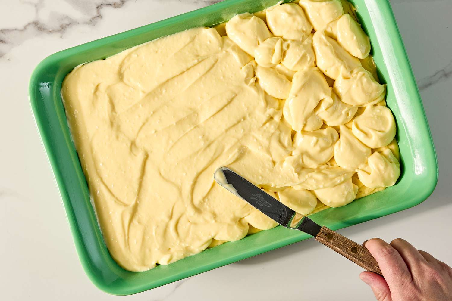 A hand spreading batter for the ugly cake recipe in a green baking tray with a spatula