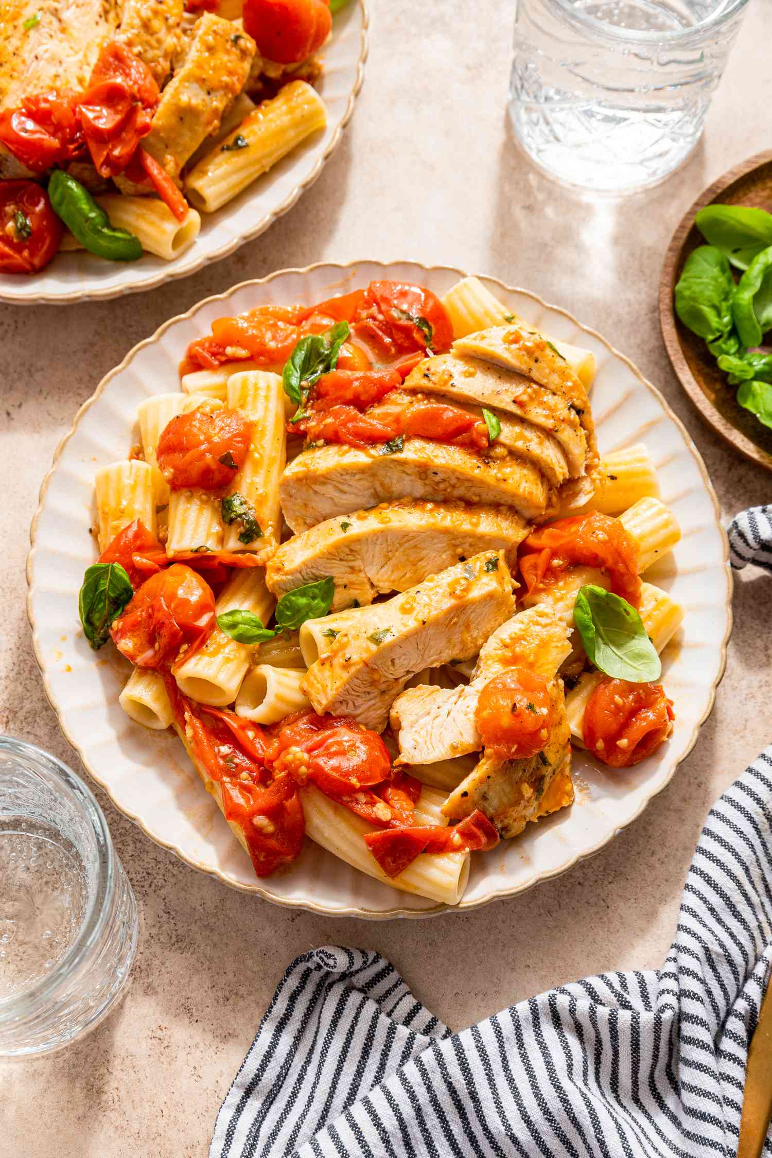 chicken pomodoro over rigatoni on a plate at a table setting with more in a cast iron skillet, glass of water, bowl of basil, and a table napkin
