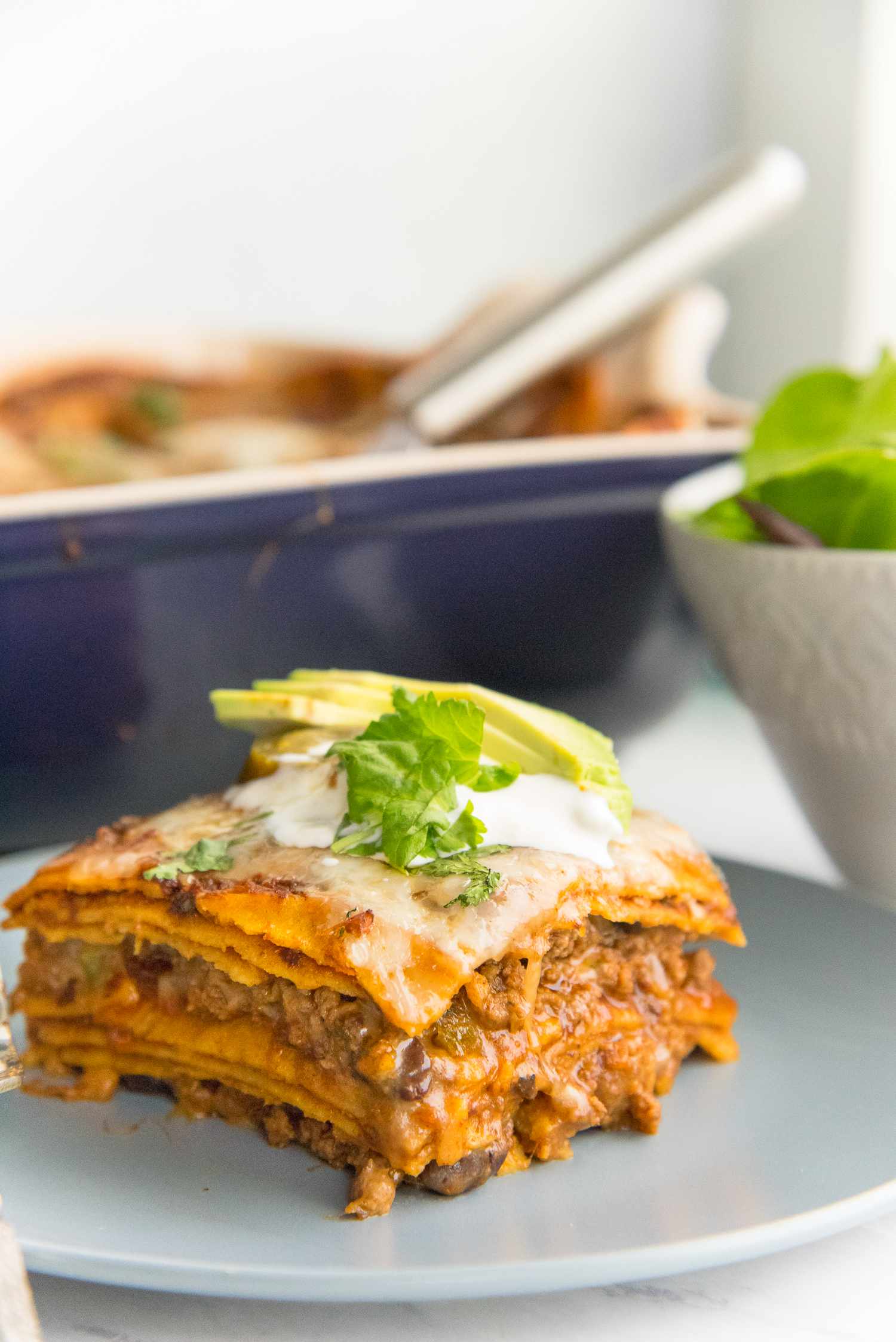 Plate with a Slice of Beef Enchilada Casserole Topped with Sour Cream, Cilantro, Jalapeno, and Sliced Avocado, and in the Background, a Bowl with Side Salad and a Casserole Dish with More