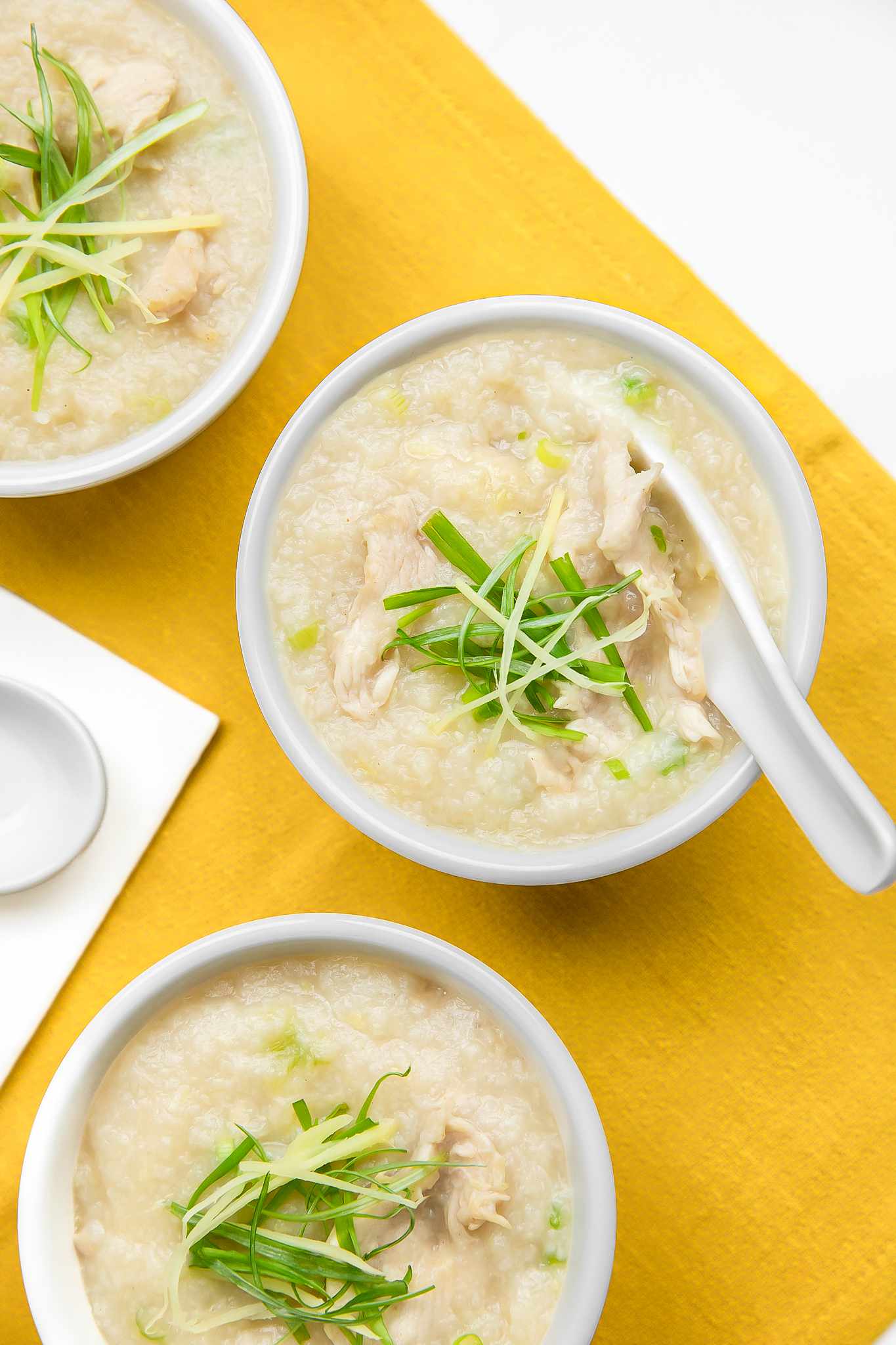 Bowls of Chicken Congee Topped with Sliced Ginger and Green Onions, Next to Soup Spoons, All on a Mustard Colored Kitchen Towel