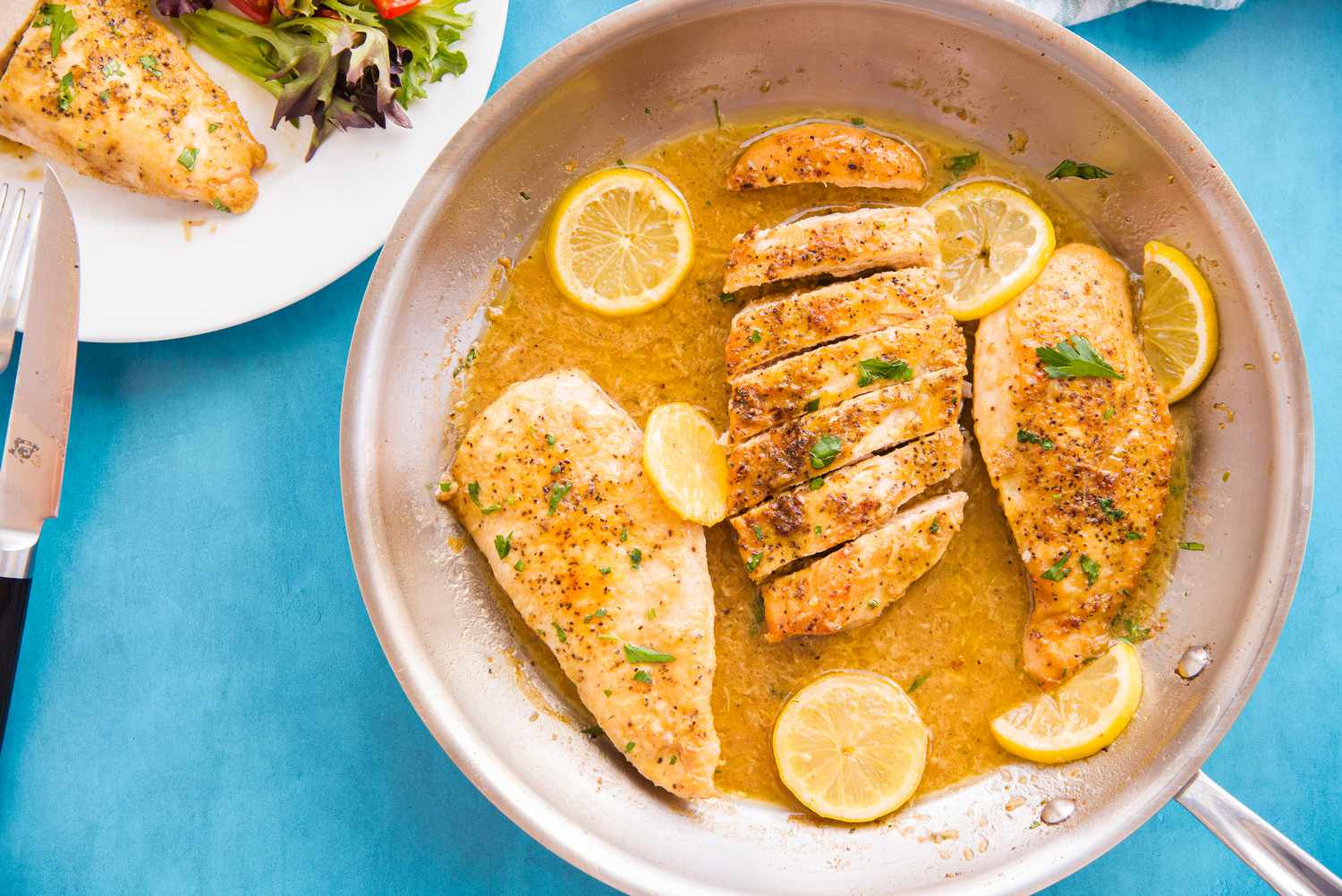 Pan of Lemon Pepper Chicken Breasts with One Sliced into Pieces and Garnished with Lemon Slices and Parsley. Next to It, a Plate with a Serving of Lemon Pepper Chicken and a Side Salad
