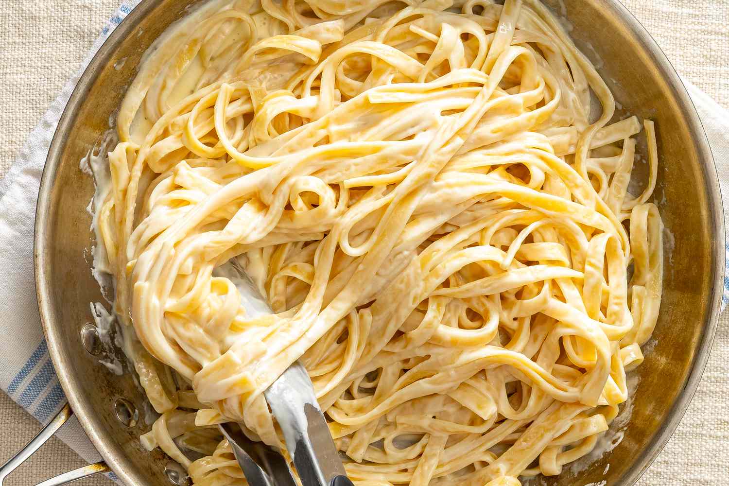 A skillet filled with fettuccine Alfredo being prepared, with pasta tongs visible