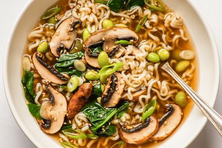 A bowl of noodle soup containing mushrooms spinach edamame green onions and sesame seeds with a spoon on the side