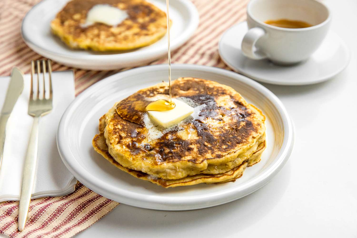 Syrup Drizzled onto a Stack of 3-Ingredient Banana Pancakes on a Plate, Surrounded by a Plate with More Pancakes, a Cup of Coffee, and Utensils on a Napkin
