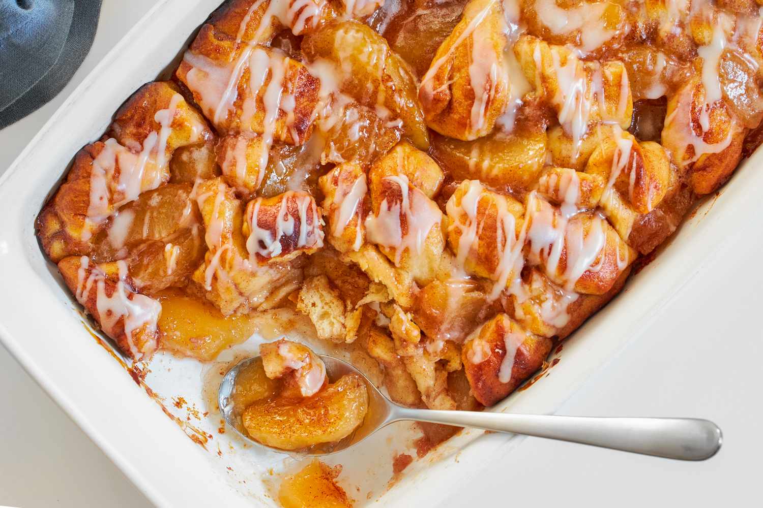 A baked apple cinnamon dish with icing in a white casserole pan a serving spoon scooping a portion from one end