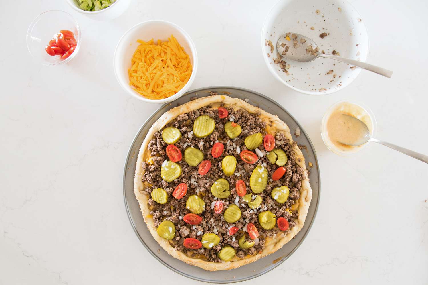 Cheeseburger Pizza Topped with Cooked Ground Beef, Pickles, and Cherry Tomatoes, and Next to It, a Bowl of Cheddar Cheese and Another Bowl with More Cherry Tomatoes