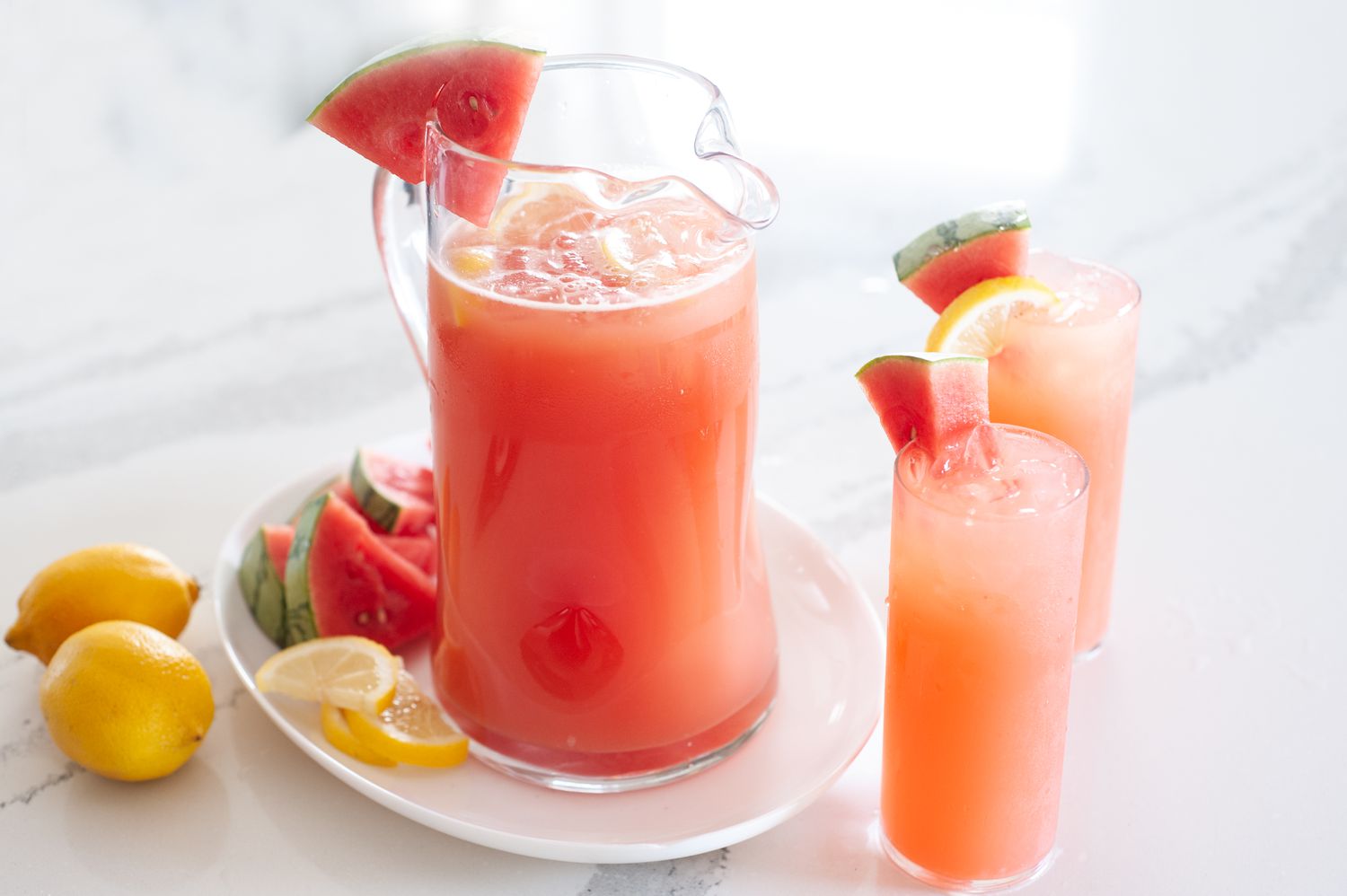 Side view of blender watermelon lemonade in a glass pitcher with two tall glasses next to it.