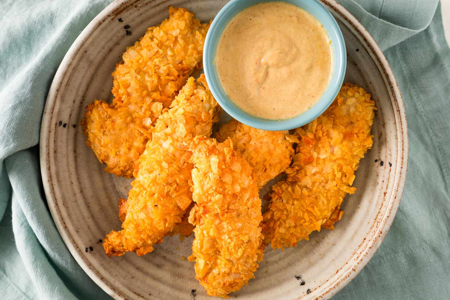 Overhead view of five air fryer potato chip chicken tenders in a stoneware bowl along with a small bowl of dipping sauce on a teal cloth background