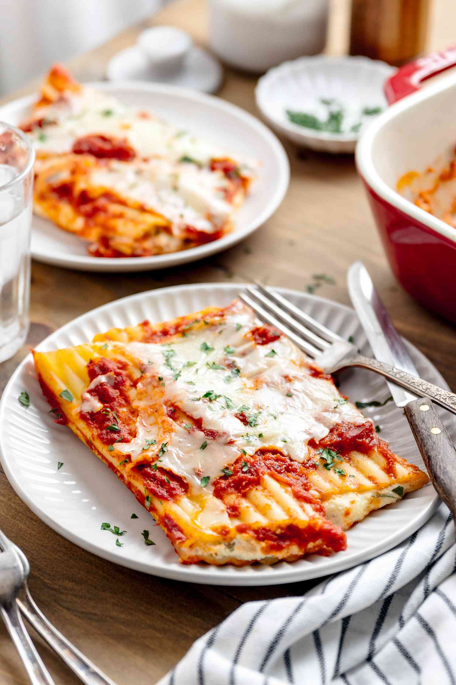 Serving of Manicotti on a Plate With Silverware, and in the Surroundings, a Table Setting With Glasses of Ice Water, a Table Napkin, Forks, Another Plate With a Serving of Manicotti, and a Casserole Dish With More Manicotti