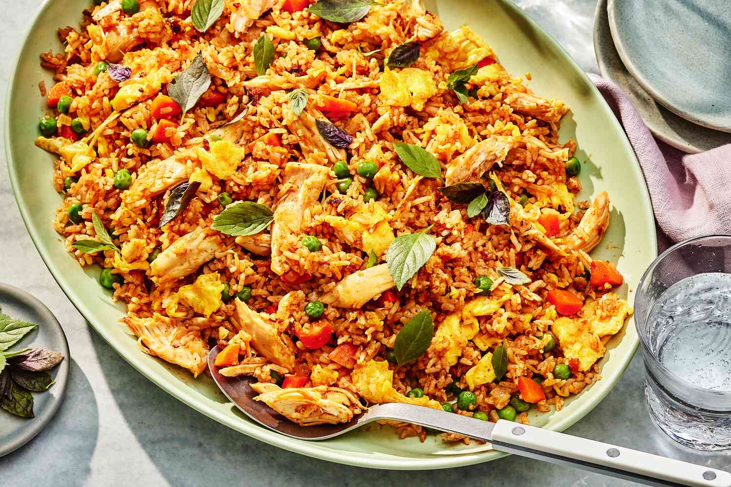 Thai-style fried rice on a plate with a serving spoon at a table setting with a stack of plates, a table napkin, a glass of water, and a small plate with Thai basil