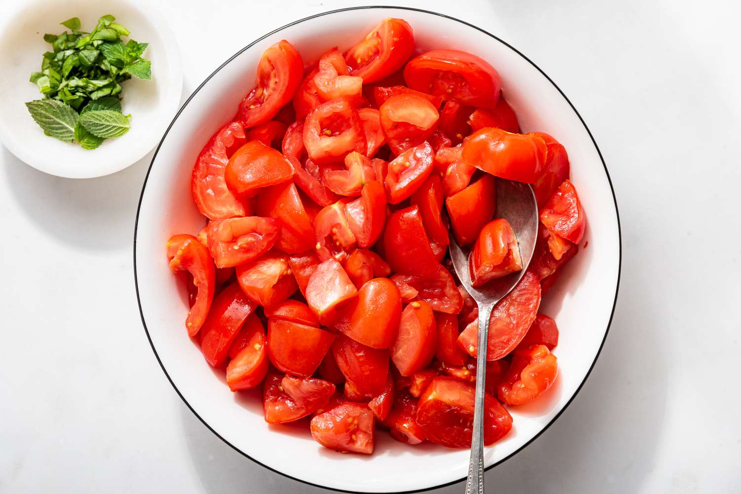 tomato quarter in a large bowl next to a smaller bowl with minced herbs for 5-ingredient tomato salad recipe