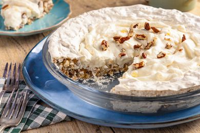 Angled view of a cream pie topped with pecans with a slice removed in a clear glass pie plate resting on a blue plate and a wooden tabletop