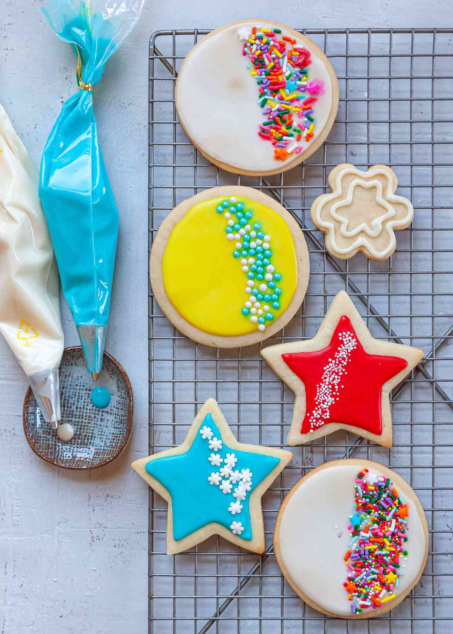 Two piping bags filled with Royal icing are set next to a cooling rack with decorated sugar cookies on it.