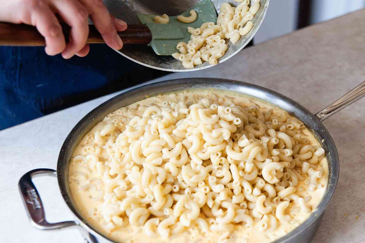 Easy mac and cheese - hand in frame holding a pot with elbow macaroni and pouring it into a pan with cheese sauce and more elbow macaroni