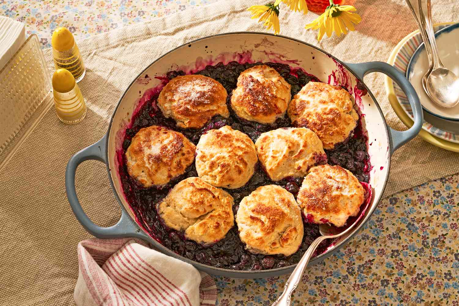blueberry grunt in a cast iron braiser at cozy looking table with a stack of bowls, salt shakers, a napkin holder with single use napkins, and a vase of flowers