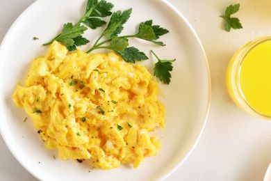Golden scrambled eggs on a plate with a sprig of parsley, next to a small glass of orange juice