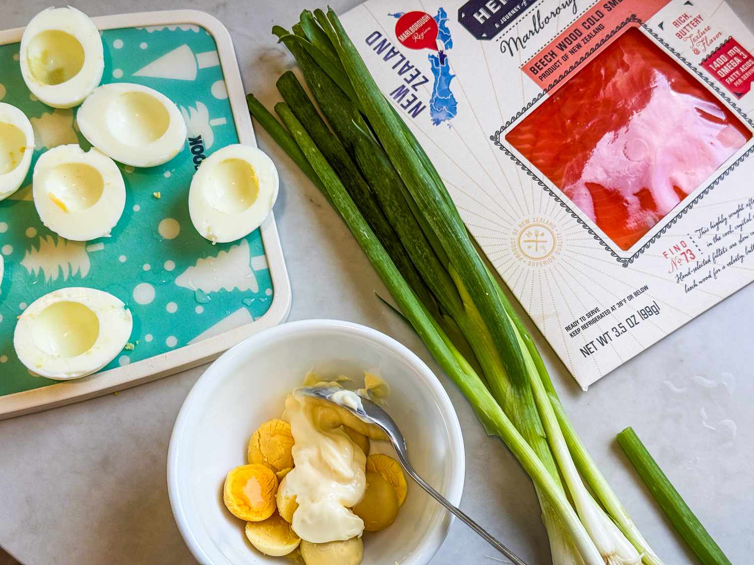 A process photo of the deviled eggs being made, showing halved hard-boiled egg whites, cooked egg yolks in a small bowl with mayonnaise, a bunch of green onions, and a package of cold-smoked salmon