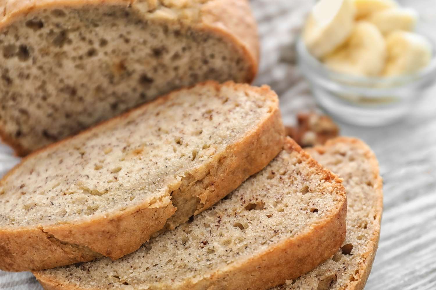 Sliced banana bread on a table with a bowl of banana slices in the background