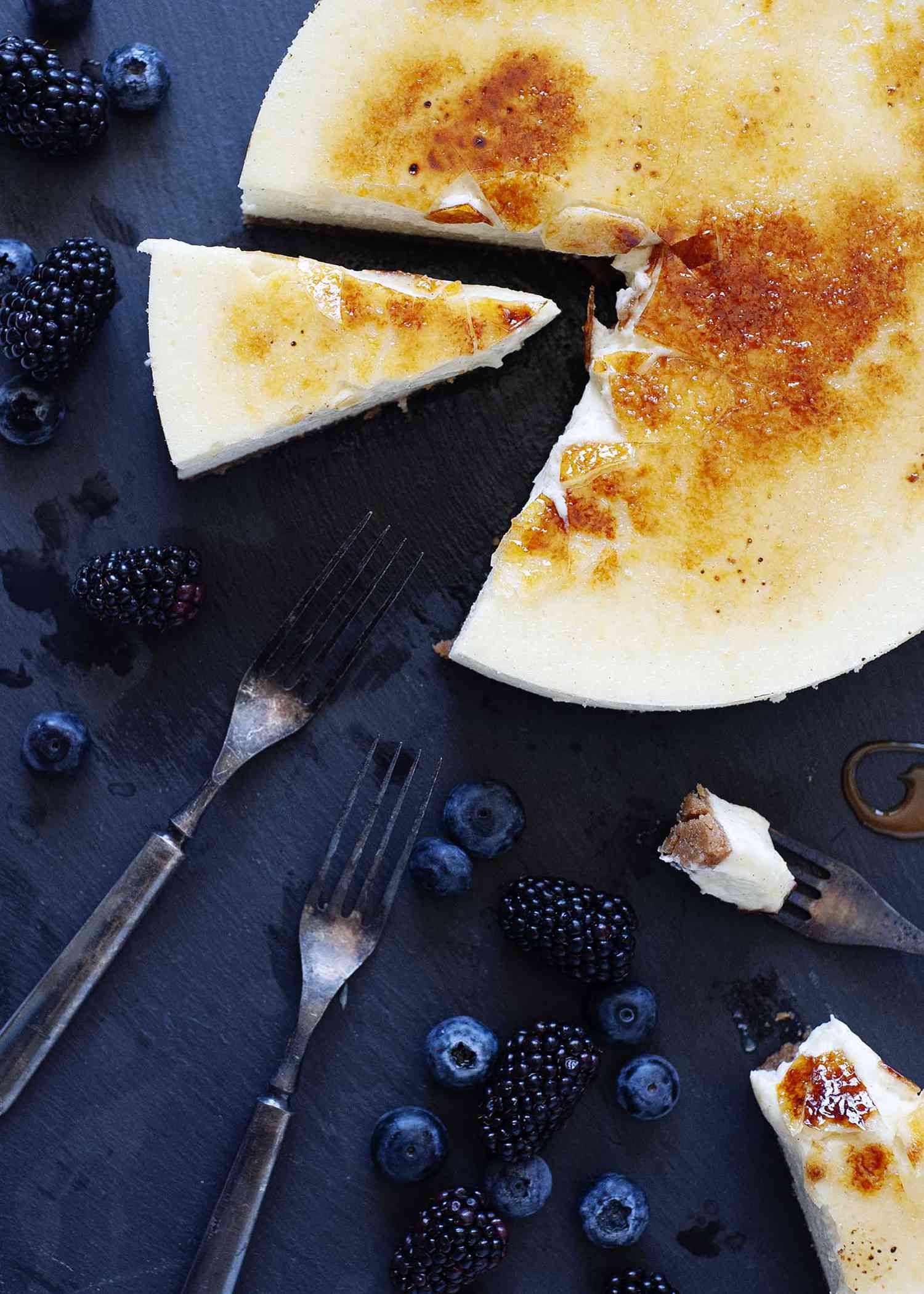Vanilla Bean Cheesecake with brittle sugar topping on a slate board wtih a slice cut out and fruit and forks nearby.