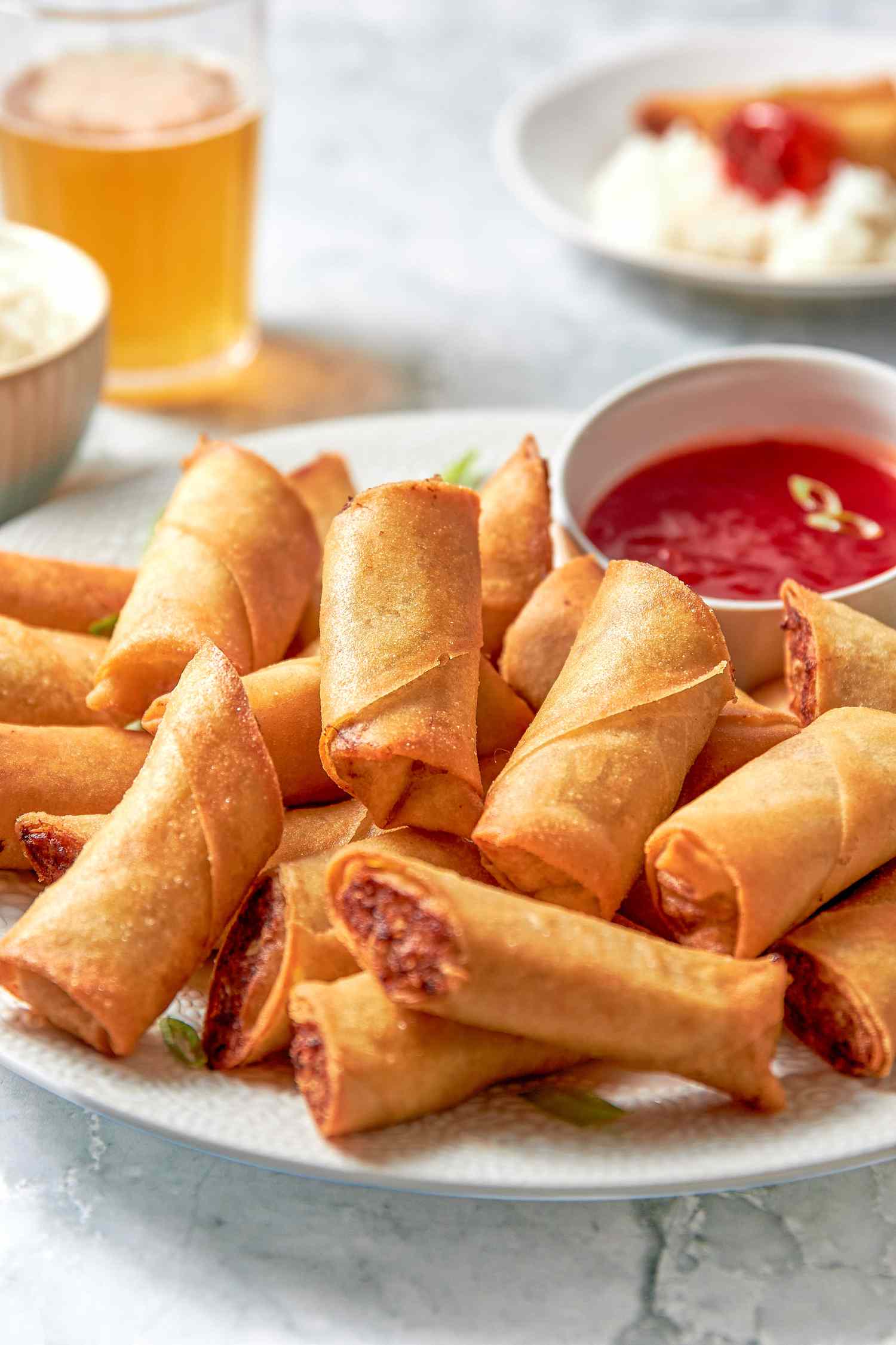 Plate of Lumpia Cut in Half With a Bowl of Sweet and Sour Sauce, and in the Background, a Bowl of Rice, a Glass With a Fizzy Drink, and a Plate With Rice, Lumpia, and Some Sauce