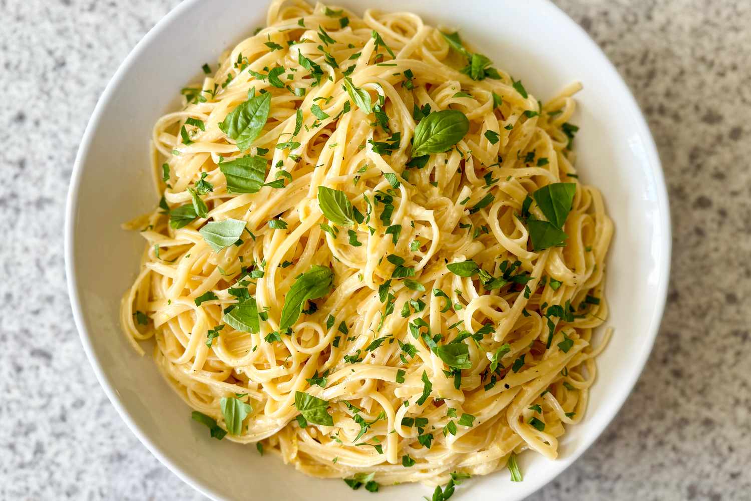 Lemon linguine topped with fresh herbs, served in a white bowl on a speckled countertop.