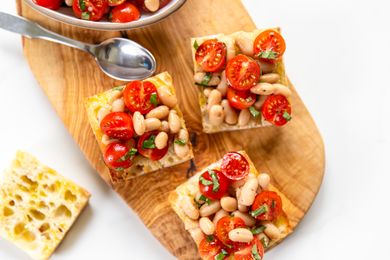 Slices of bread topped with cherry tomatoes white beans and herbs on a wooden serving board with a spoon visible nearby