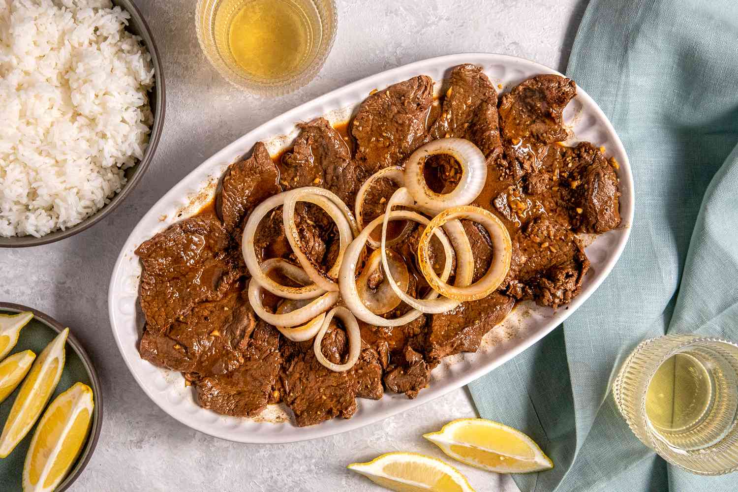 Plate of Bistek Topped With Onions Next to a Two Glasses, Lemon Wedge on the Counter and More in a Bowl, a Bowl of Rice, and a Light Blue Table Napkin