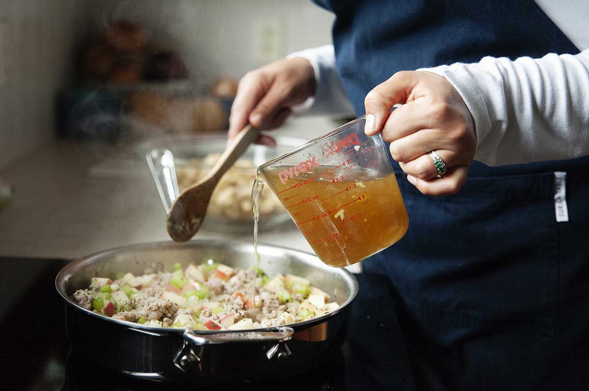 Woman pouring stock into a pan full of pork sausage, celery and sage for easy, homemade stuffing.