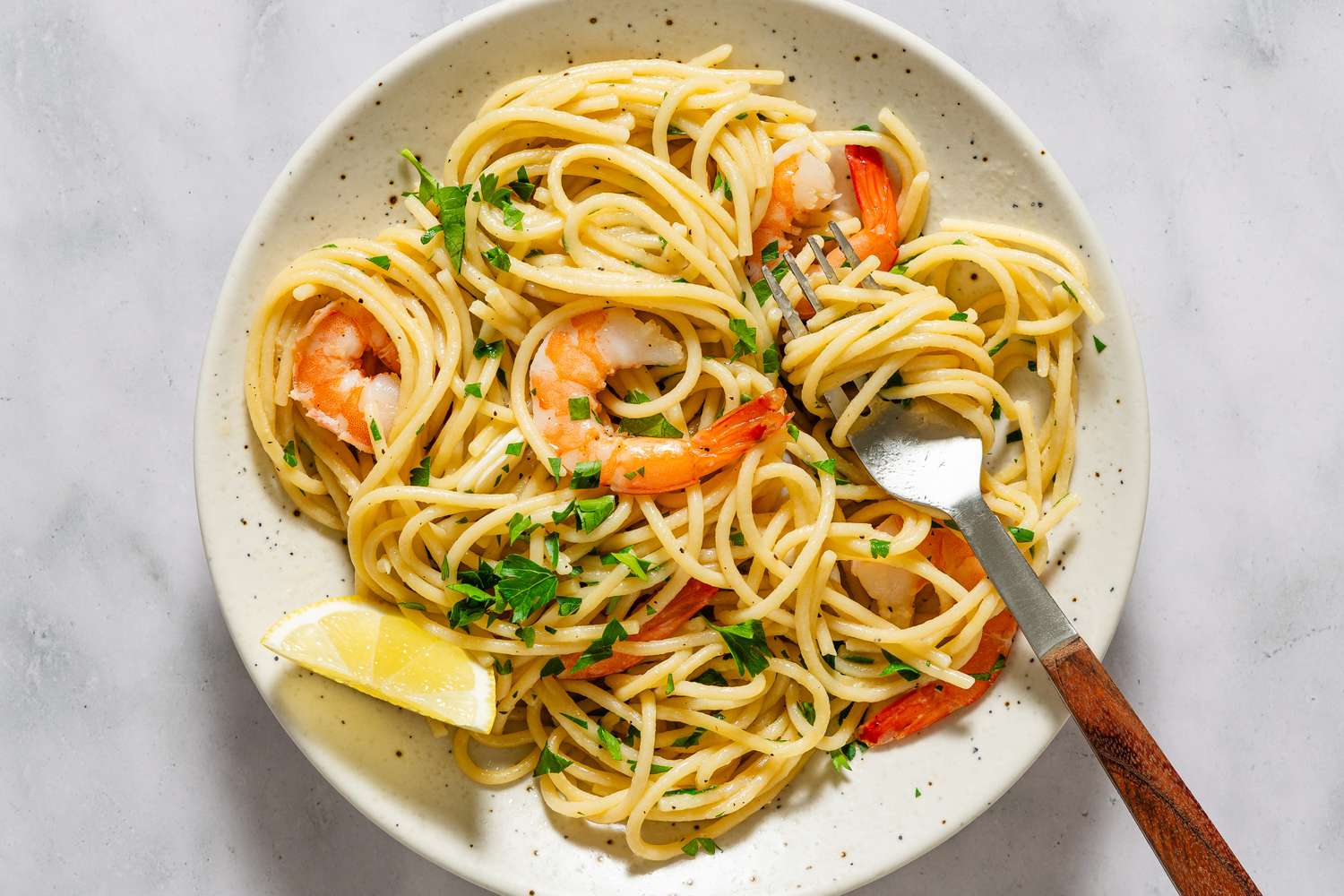 A plate of spaghetti with shrimp garnished with herbs served with a lemon wedge on the side fork resting on the plate