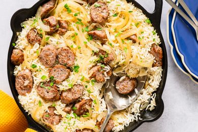 A skillet containing a sausage and rice bake with onions and fresh herbs a serving spoon resting inside