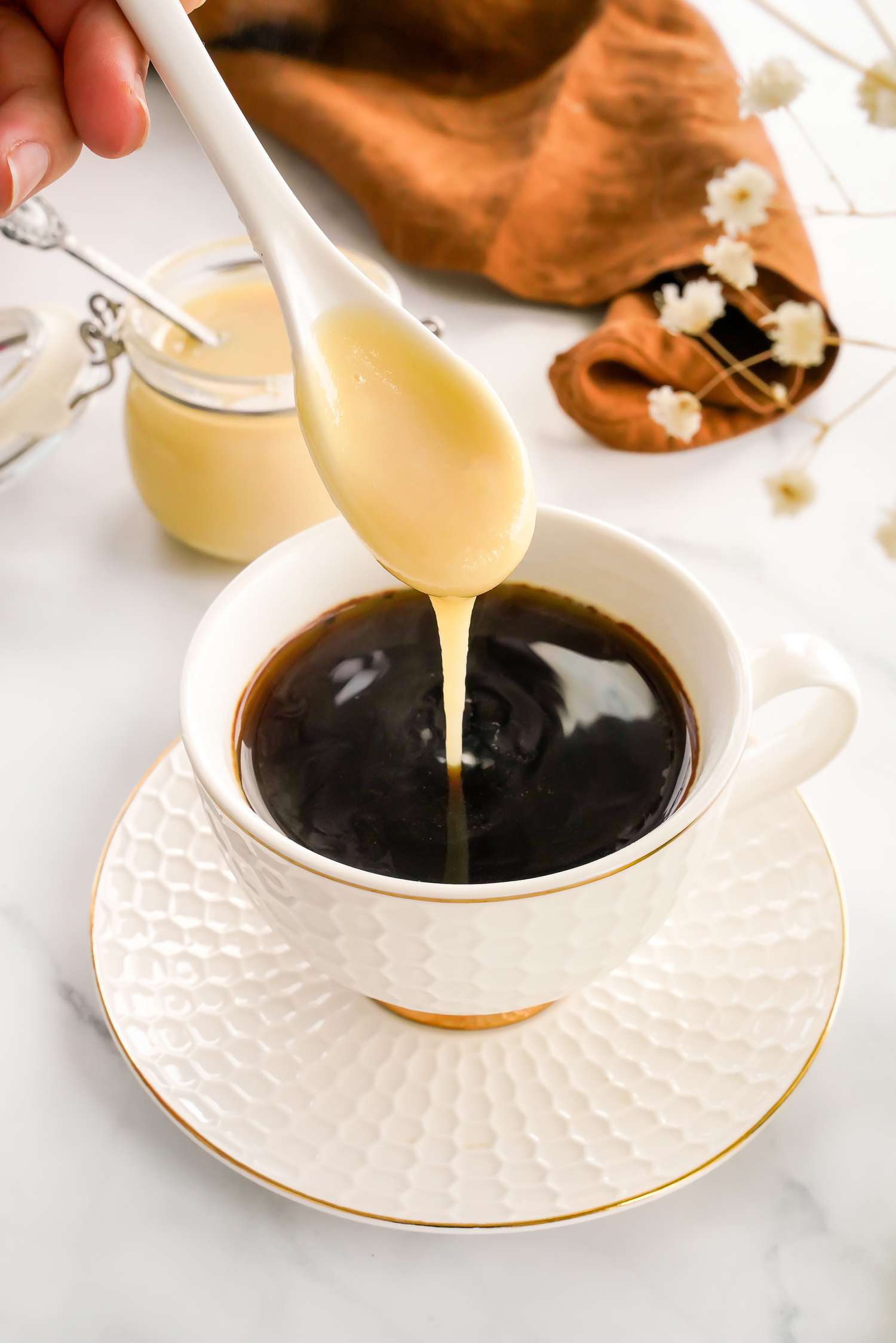 Spoonful of Homemade Condensed Milk Poured Into a Cup of Coffee, and in the Background, a Kitchen Towel and a Jar With More Condensed Milk