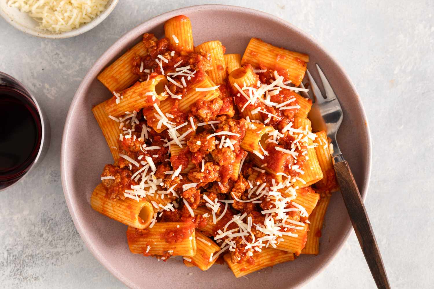 A bowl of pasta with sausage sauce and grated cheese a fork on the plate