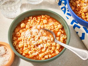 Overhead view of a green bowl of Pasta e Ceci and a spoon topped with parmesan cheese next to a bowl of parmesan, another bowl of pasta and a glass of carbonated beverage