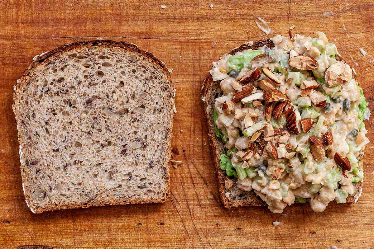 Chickpea salad is on top of a slice of bread and topped wth chopped almonds. A slice of sandwich bread is to the left.