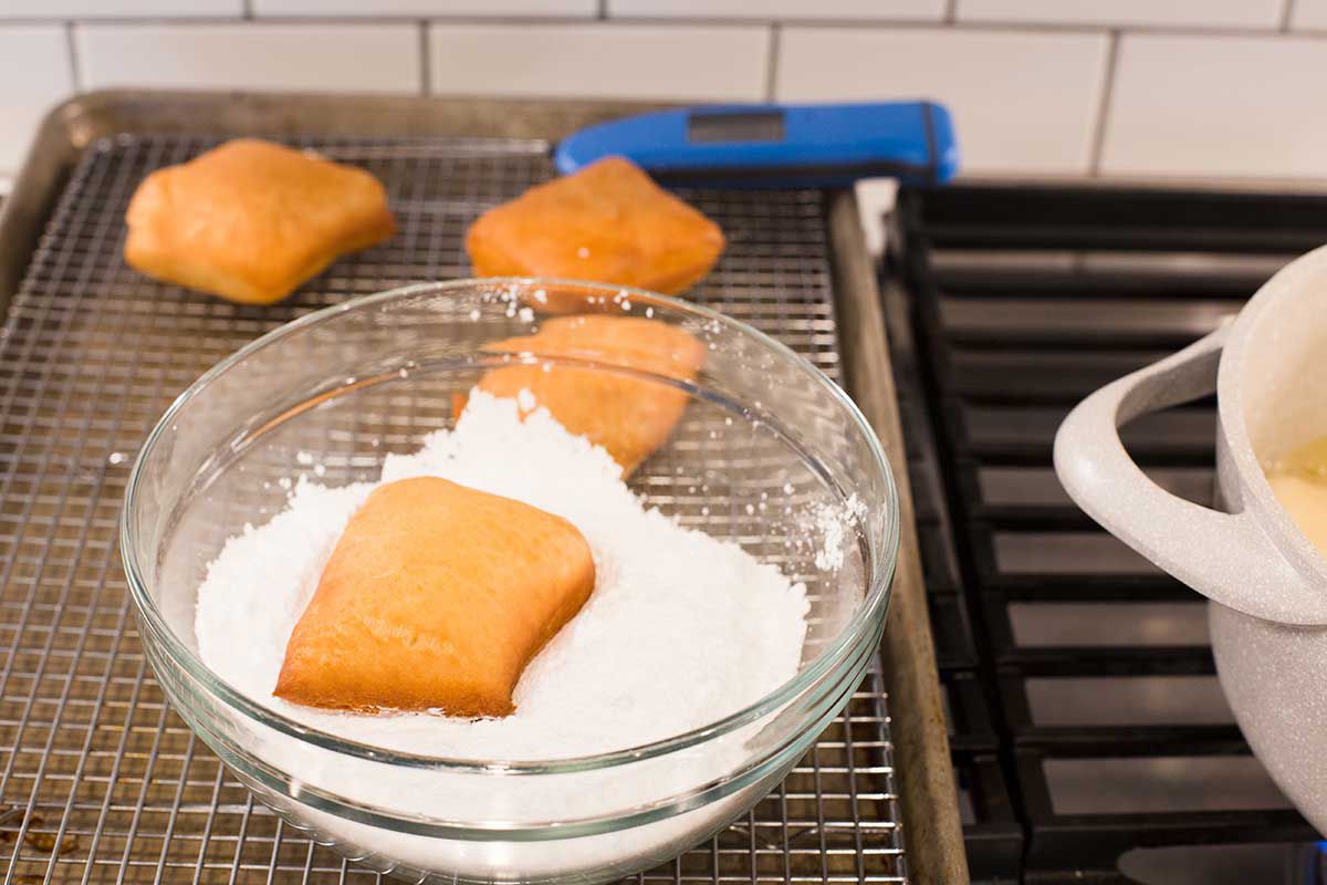 New Orleans Beignets that have been fried. One is in a bowl of powdered sugar and three more are behind it on a baking tray. A partial view of a dutch oven on a stove is to the right.