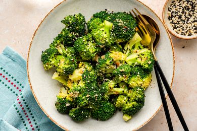 Bowl of Microwave Broccoli with Sesame Dressing with Utensils, Next to a Table Napkin, a Glass of Water, and a Bowl with More Sesame Seeds