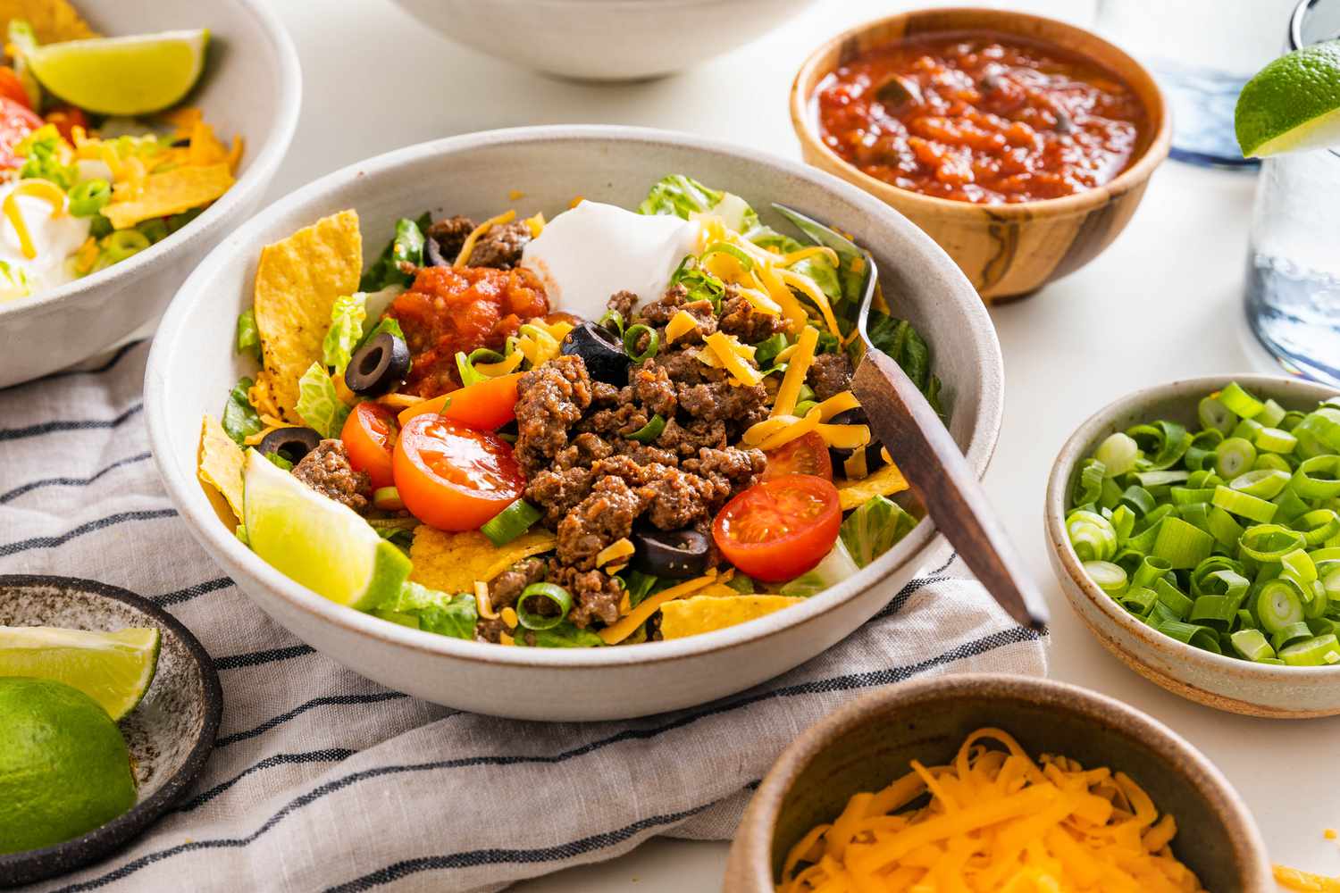 Bowl of Taco Salad Surrounded by Another Serving in a Bowl, a Bowl of Salsa, a Bowl of Sliced Scallions, a Bowl of Shredded Cheese, and a Bowl With Quartered Limes